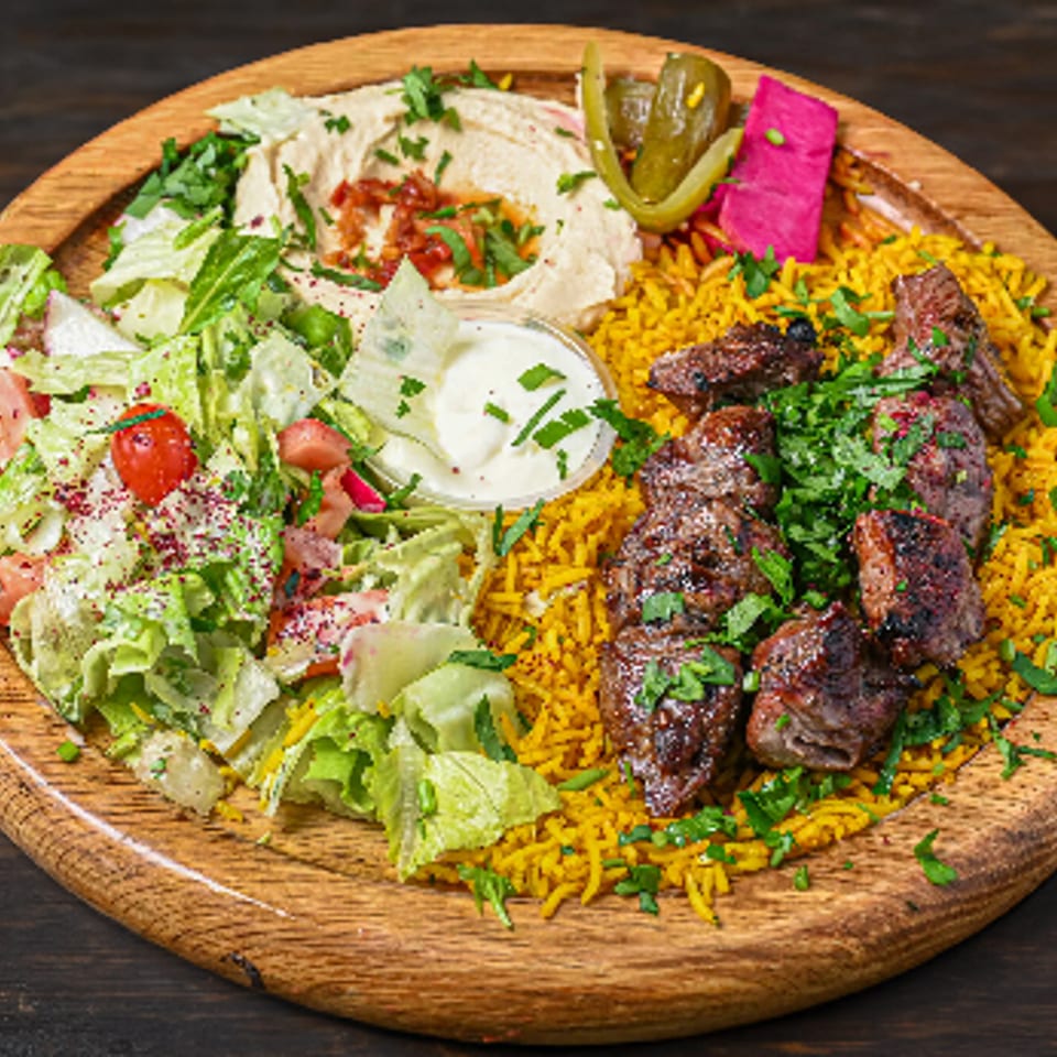 Middle Eastern platter with grilled meat, yellow rice, fresh salad, hummus, and pickled vegetables on a wooden board.