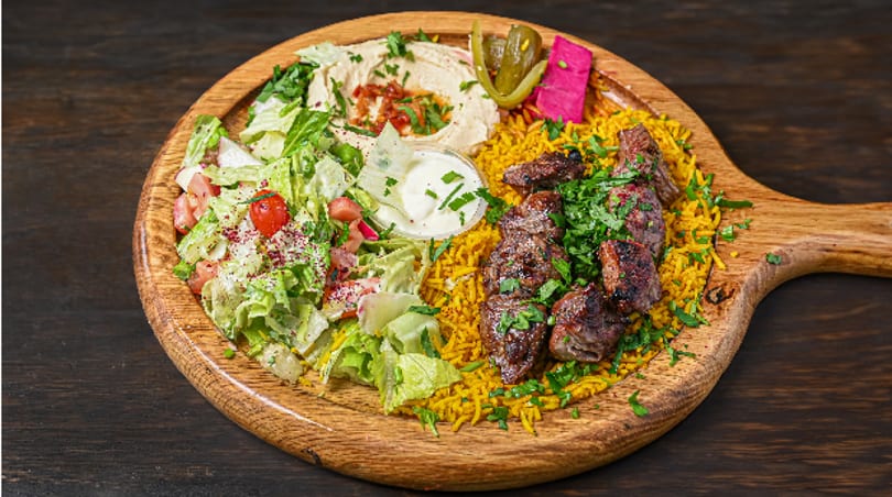 Middle Eastern platter with grilled meat, yellow rice, fresh salad, hummus, and pickled vegetables on a wooden board.