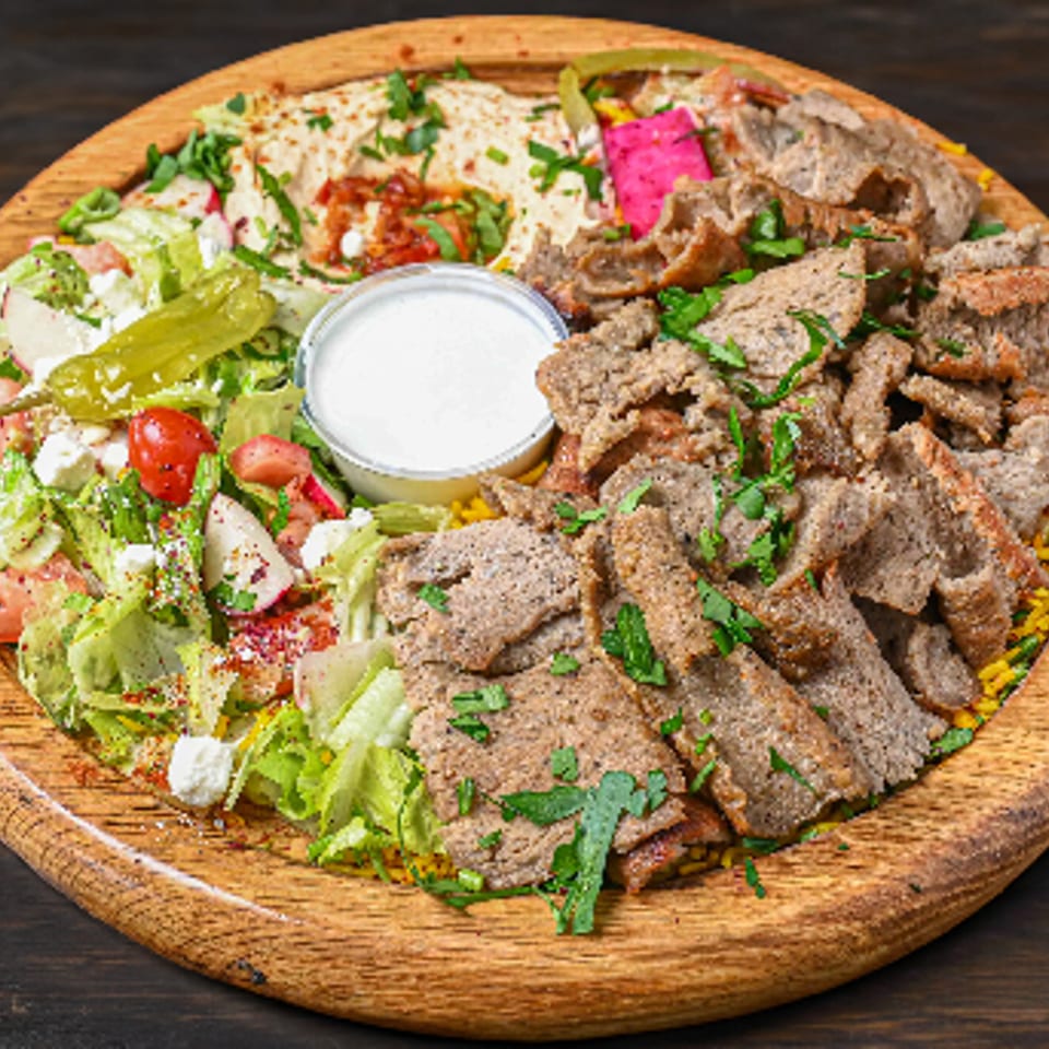 Gyro meat platter with fresh salad, hummus, rice, and white sauce on a wooden board.