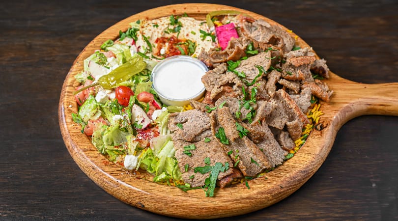 Gyro meat platter with fresh salad, hummus, rice, and white sauce on a wooden board.