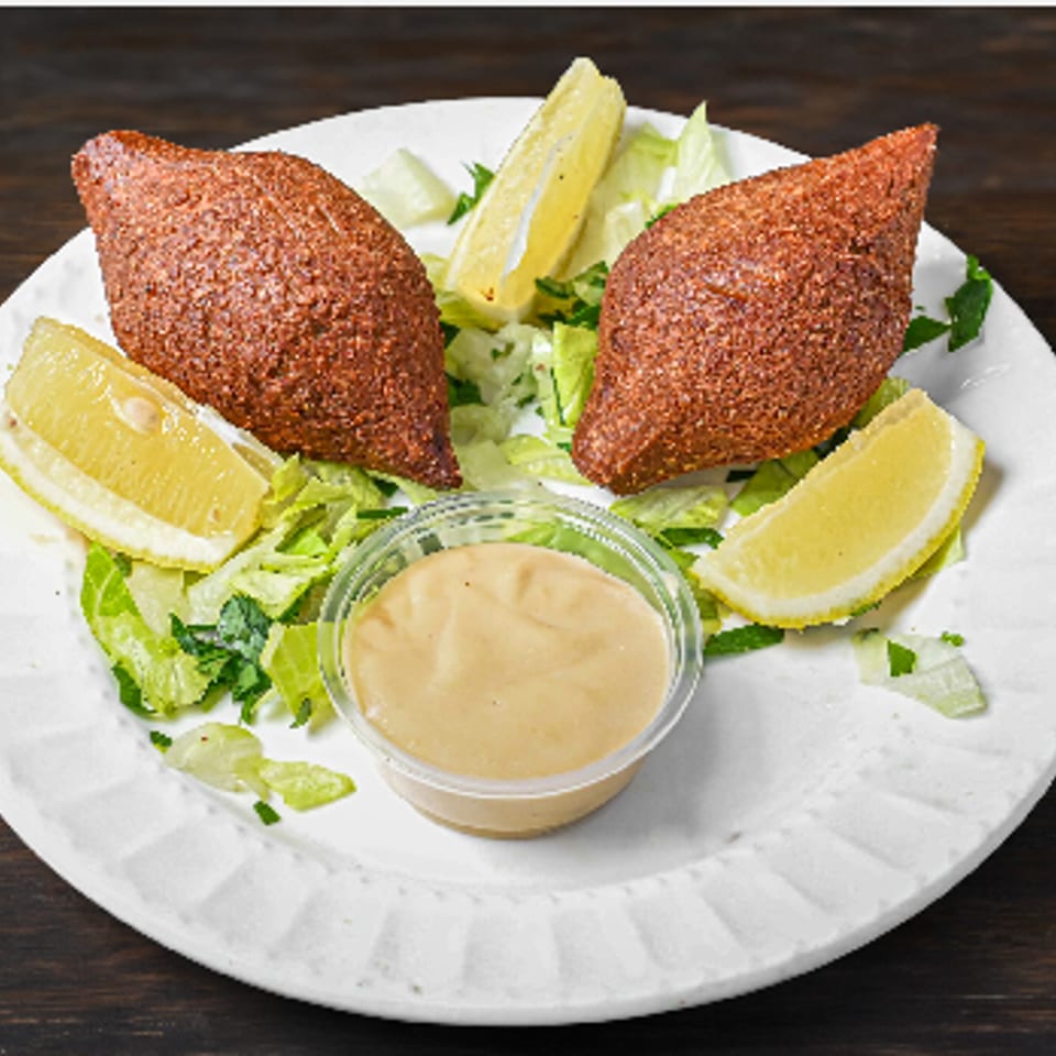 Two fried kibbeh on a white plate with lettuce, lemon wedges, and a dipping sauce.