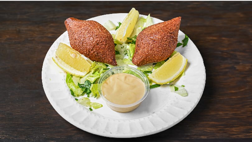 Two fried kibbeh on a white plate with lettuce, lemon wedges, and a dipping sauce.